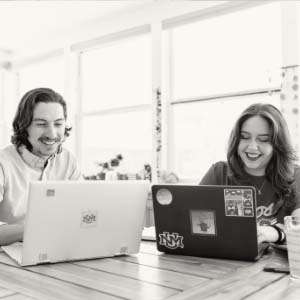 Two people sitting at a table looking at their laptops and smiling